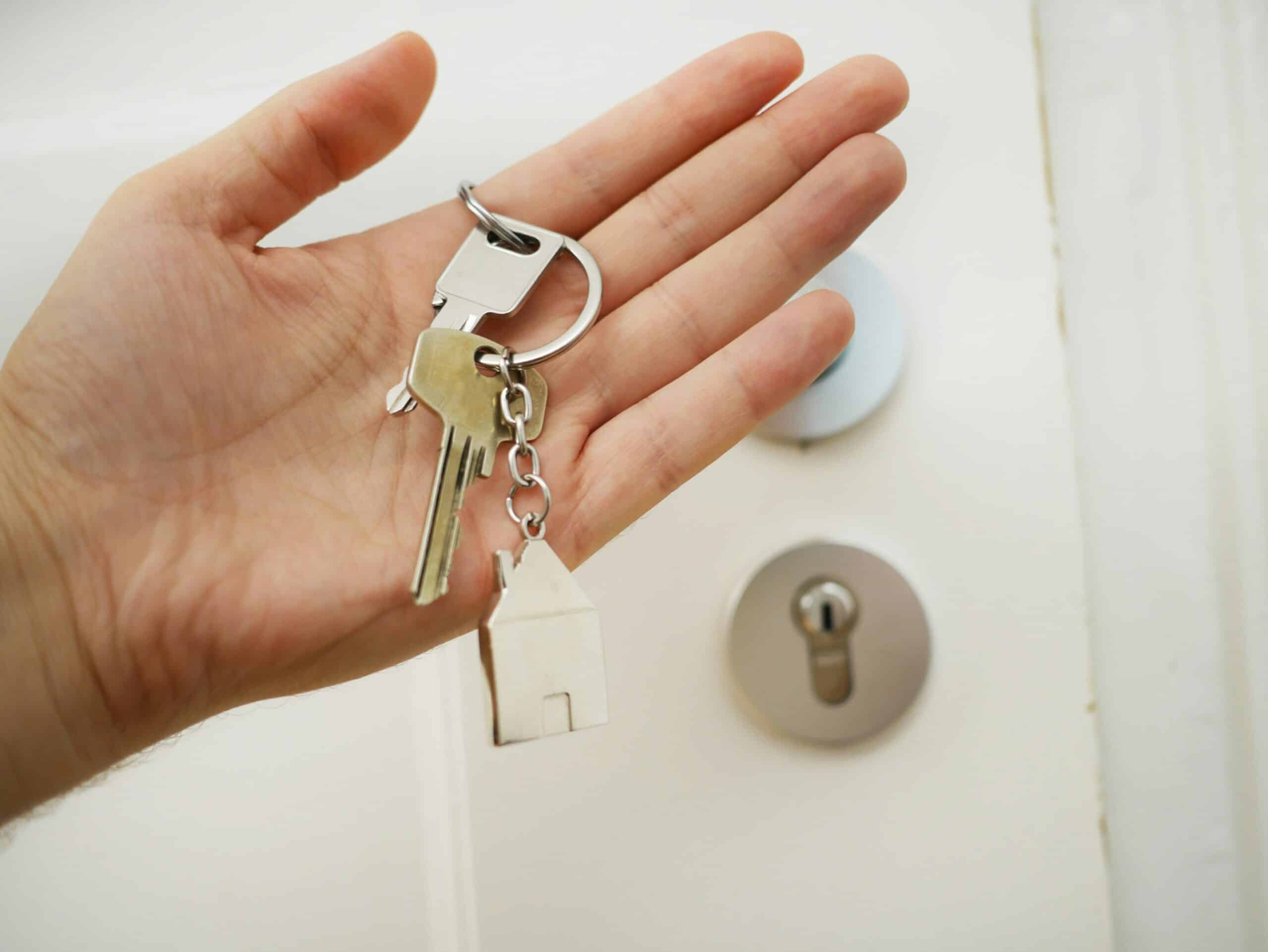 Close view of a hand holding house keys; the front door of a house in the background.